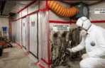 Professional Technician Executing Mold Remediation Process Steps in Basement A photo illustrating several mold remediation process steps. A technician in a Hazmat suit inspects a black moldy wall with a moisture meter. Behind him, a containment zone is sealed with plastic, and a HEPA air scrubber is running, showing the initial setup for professional mold removal.