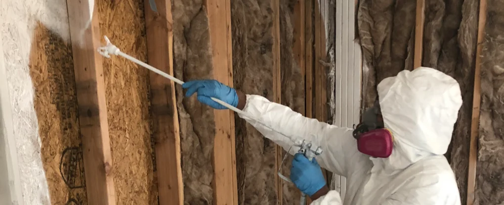 A specialist in a full hazmat suit and respirator applying antimicrobial spray to attic insulation and rafters during attic mold remediation in Charlotte NC.