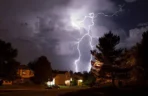 How Sudden Summer Thunderstorms Cause Hidden Crawl Space Mold Bright lightning bolts striking over a residential suburban neighborhood during a intense night storm, bringing heavy rainfall and high humidity.