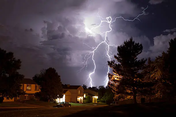 Bright lightning bolts striking over a residential suburban neighborhood during a intense night storm, bringing heavy rainfall and high humidity.