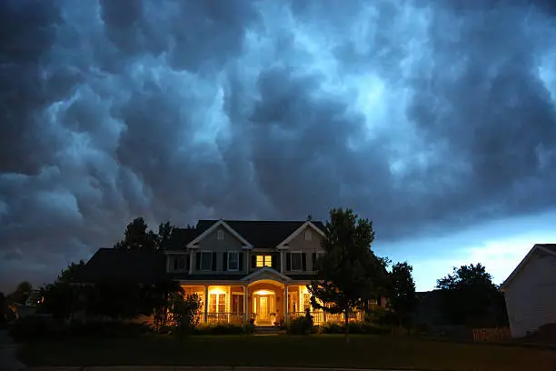 Heavy, dark mammatus rain clouds gathering over a lit two-story suburban house right before a severe regional summer downpour.