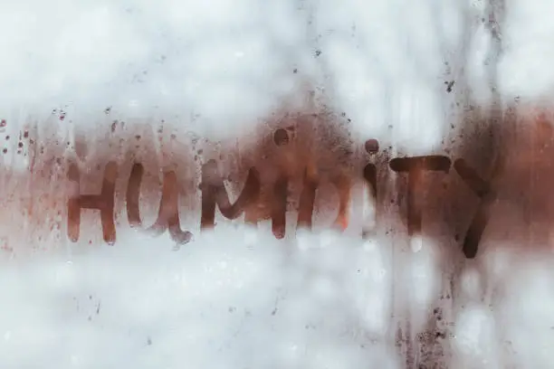 The word "HUMIDITY" written in condensation on a heavily fogged glass window, demonstrating why Charlotte's summer humidity is a breeding ground for attic mold.