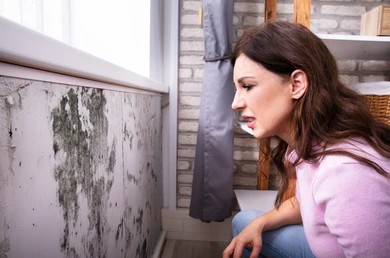 A concerned woman inspecting dark mold growth on a interior wall near a window..