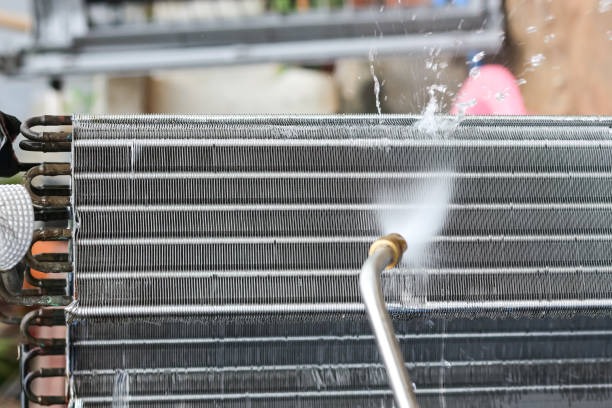 Pressure washer wand spraying high-pressure water to deep clean metal cooling coil fins on an HVAC evaporator assembly.