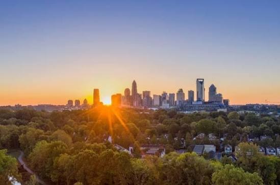 Panoramic aerial view of the Charlotte North Carolina skyline at sunset with high sun glare and surrounding tree canopy.
